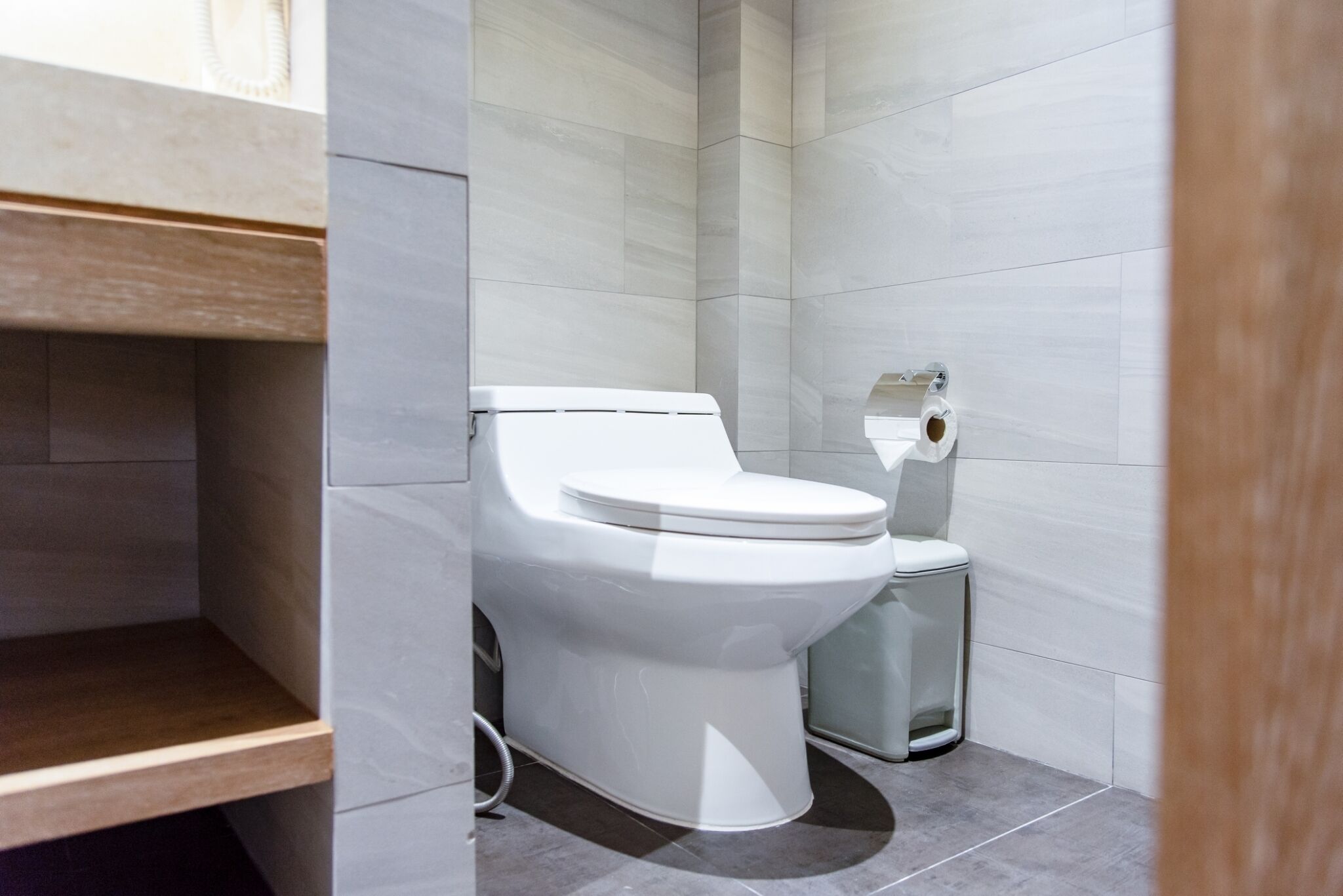 A modern bathroom with a white toilet, a trash bin, and a wooden shelf.