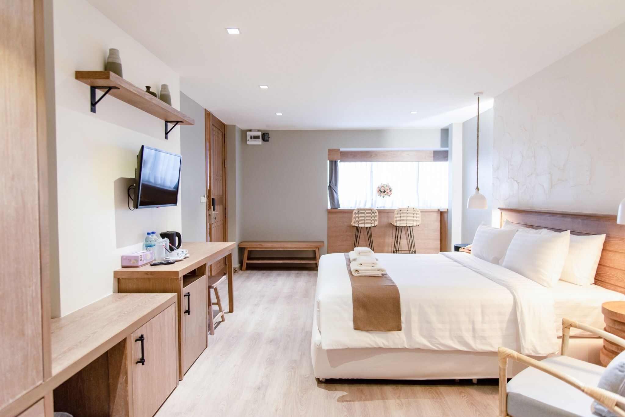 Una habitación de hotel moderna con una cama grande, un escritorio con televisor de pantalla plana, un banco de madera y una pequeña mesa con un hervidor.