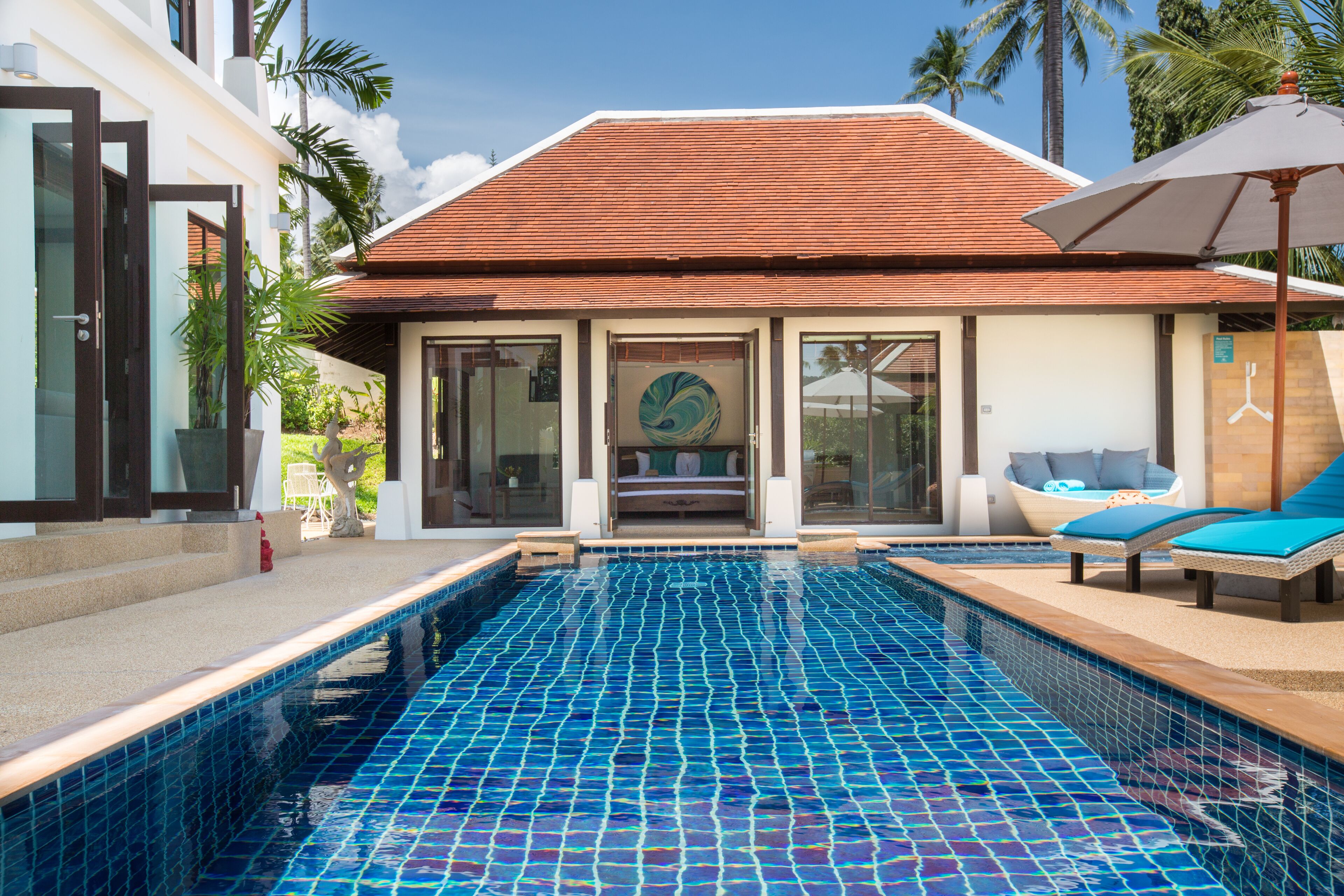 A swimming pool with a tiled floor, a building with a tiled roof, and a lounge area with blue cushions.