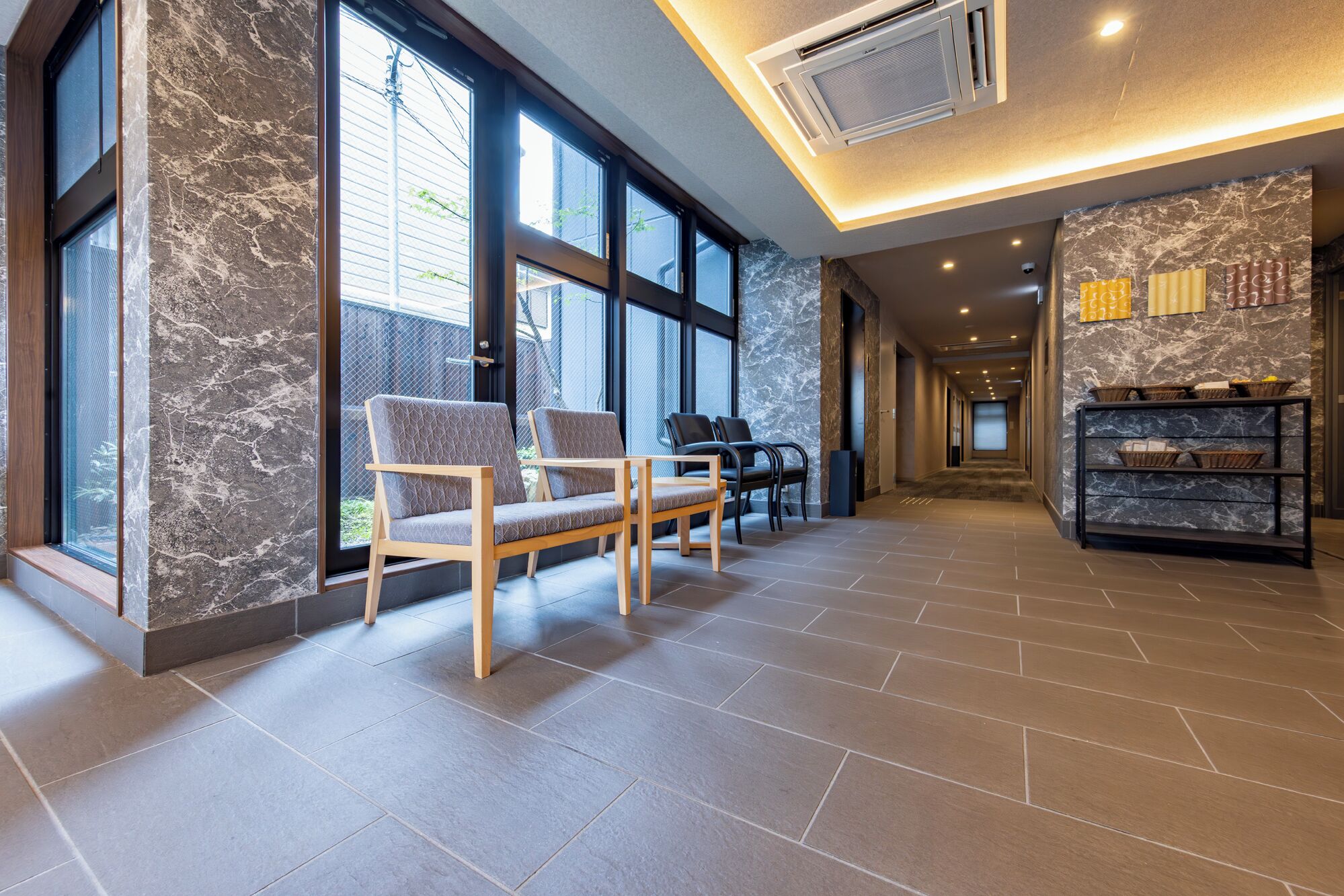 A spacious lobby with large windows, a seating area with chairs, a reception desk, and a marble wall.