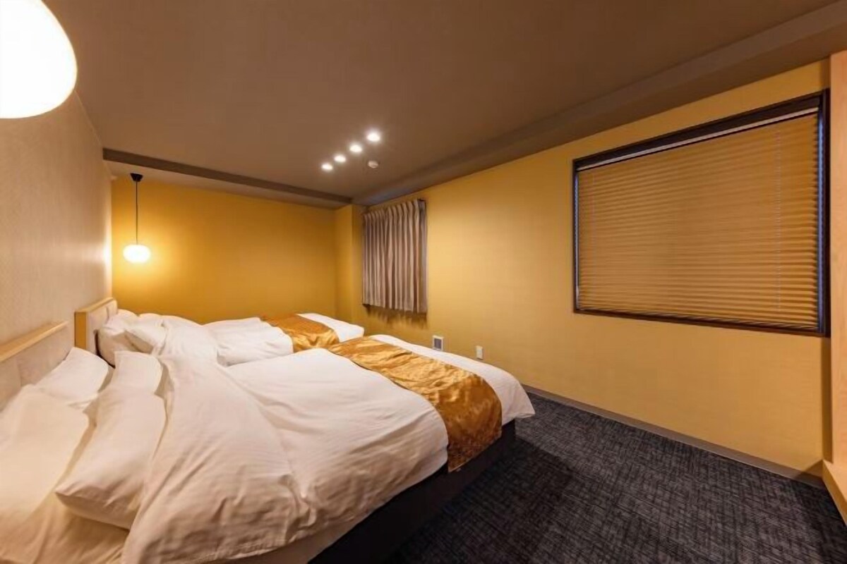Une chambre d’hôtel avec deux lits, une grande fenêtre avec stores, un luminaire fixé au mur et un sol recouvert de moquette.