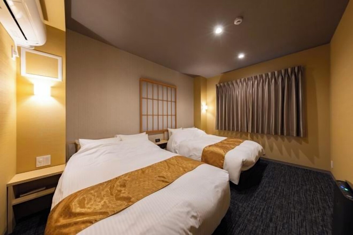 A hotel room with two beds, a bedside table, a wall-mounted air conditioner, a window with curtains, and a wall-mounted light fixture.