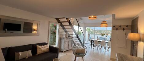Un salon moderne avec un canapé, une table basse et un coin repas avec des chaises. Un escalier mène à une terrasse extérieure offrant une vue dégagée.