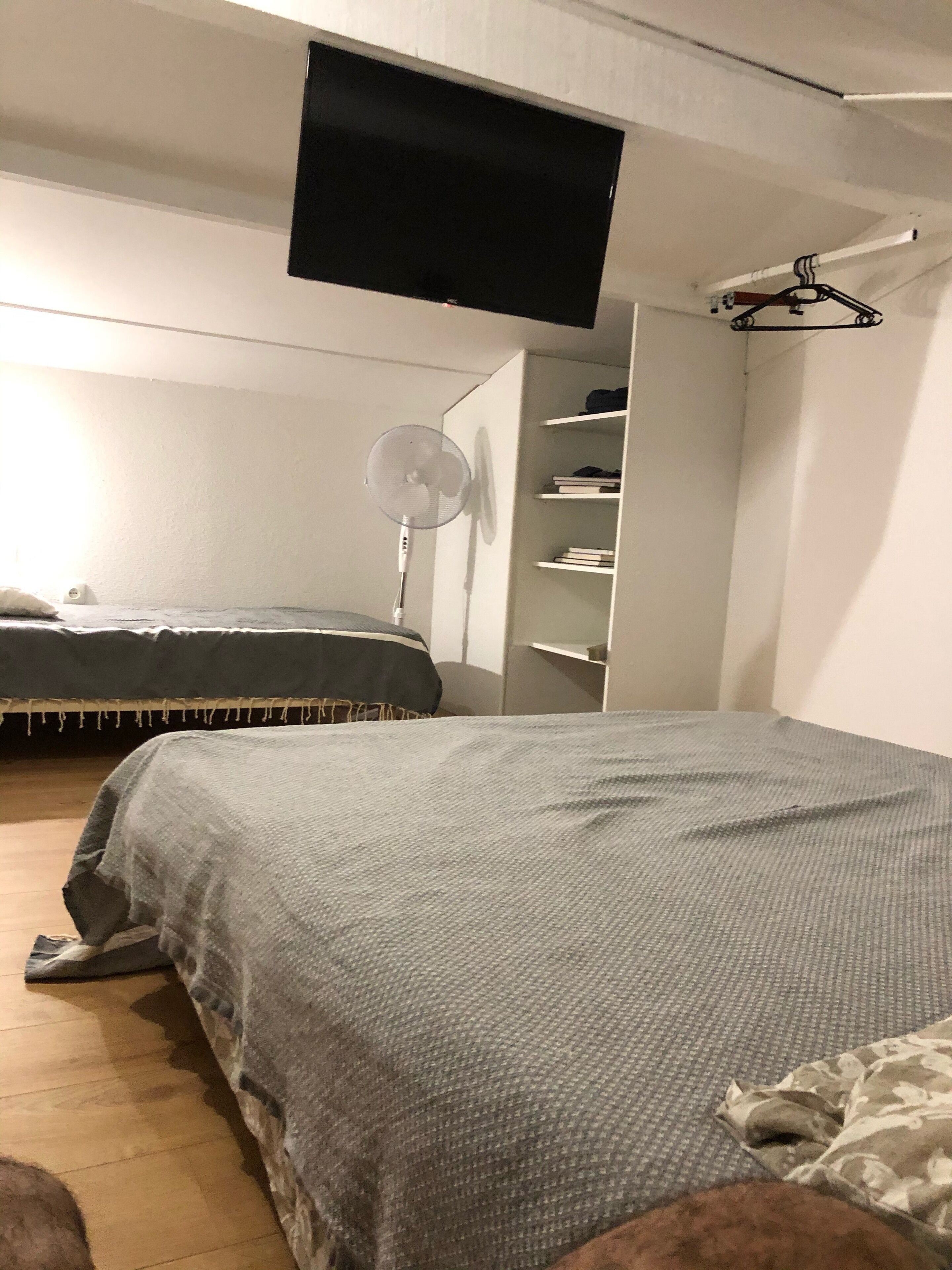 A bedroom with a bed, a TV mounted on the wall, a fan, and a shelving unit.