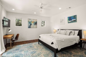 Quarto com cama, escrivaninha, cadeira, ventilador de teto e obras de arte nas paredes.