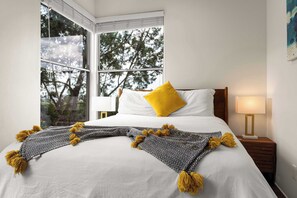 A hotel room with a large window, a bed with white bedding and a yellow pillow, a patterned throw blanket, and a bedside lamp.