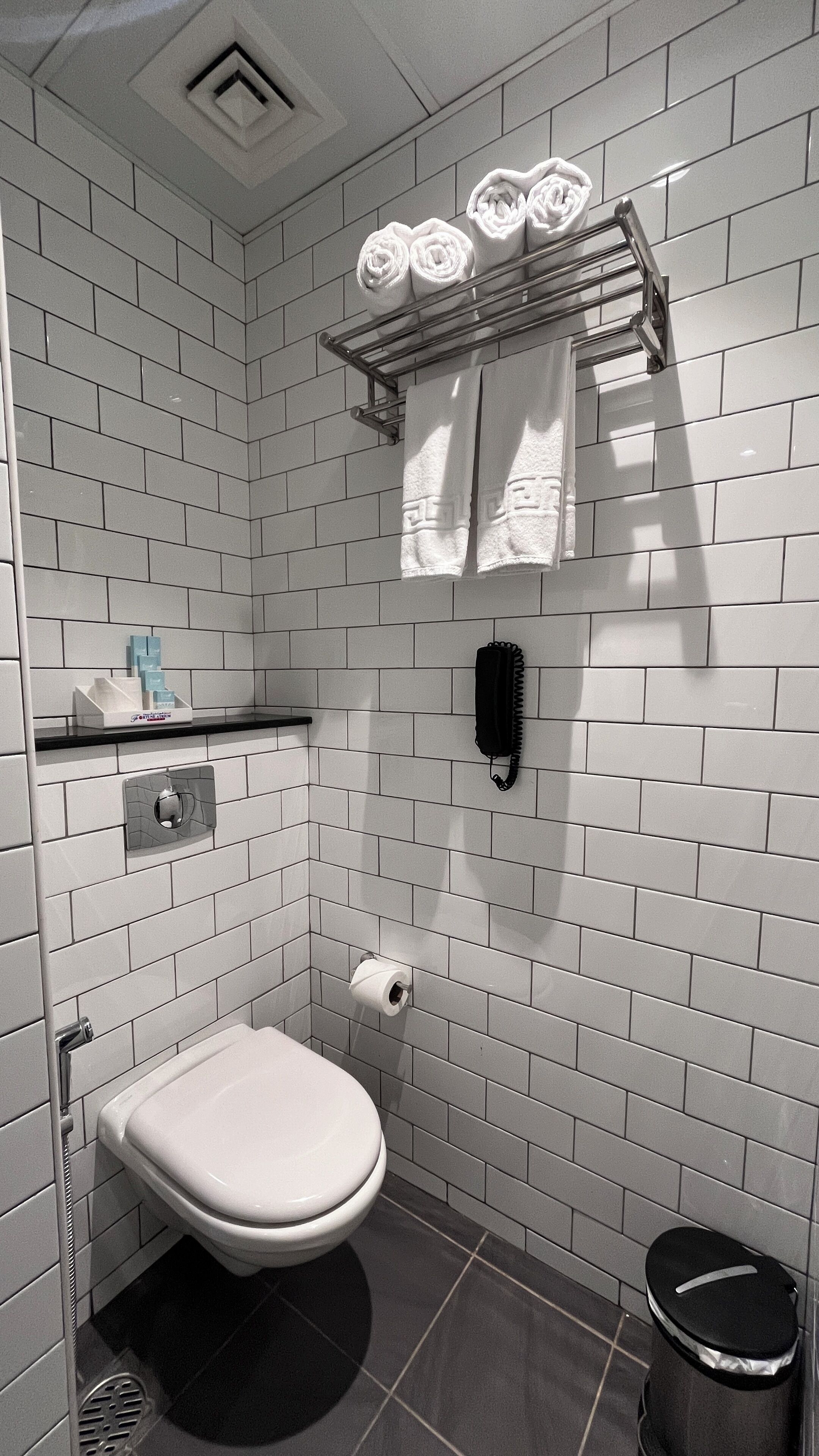 A modern bathroom with white subway tiles, a white toilet, a black trash bin, a towel rack with rolled towels, a black telephone, and a soap dispenser on a shelf.