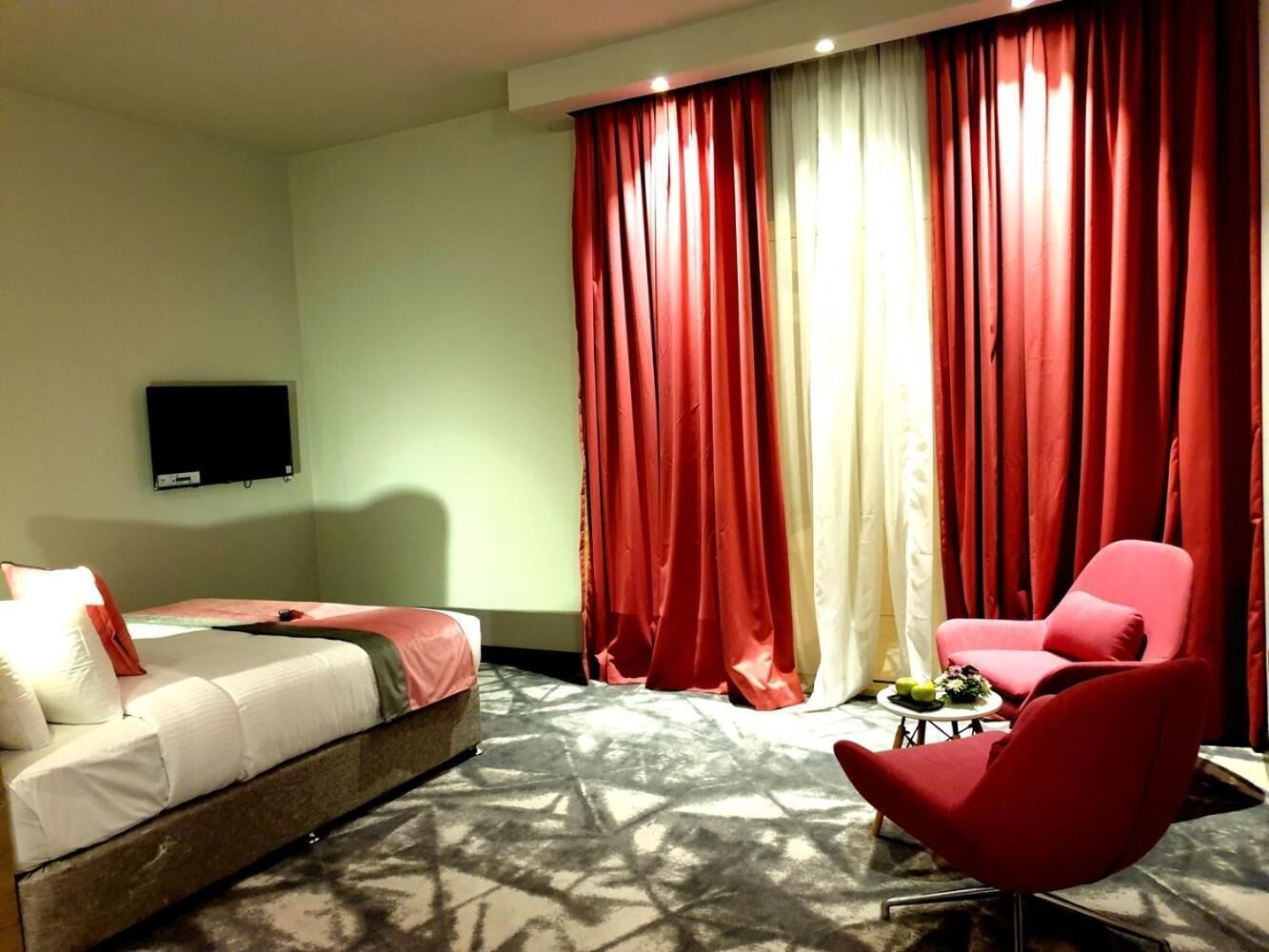 A hotel room with a bed, a red armchair, a small table with a fruit bowl, and red curtains.
