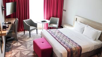 A hotel room with a bed, two chairs, a small table, a TV, and a desk with a computer and a lamp.
