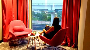 Una habitación con un ventanal grande, cortinas rojas, una silla roja y una pequeña mesa con frutas.