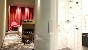 Habitación de hotel con una cama, una silla roja, una mesita con frutas, un armario y un baño con ducha y lavabo.
