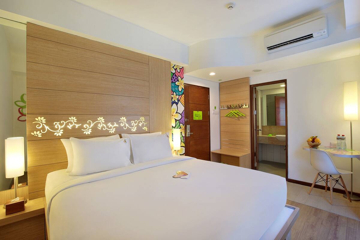 Habitación de hotel con una cama grande, una mesita de noche con lámpara, un pequeño escritorio con silla, y un baño visible a través de una puerta abierta.