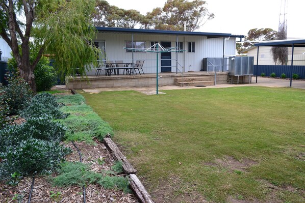 A residential backyard with a lawn, a patio with outdoor furniture, a clothesline, and a building with a blue roof.