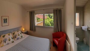 A bedroom with a bed, a nightstand, a red chair, a window with curtains, and a view of a deck. - Apartment with terrace overlooking the park (Clohars Fouesnant)