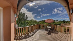 A wooden deck with a table and chairs, a view of trees and a clear sky, and an indoor living area with a sofa and a TV. - Apartment with terrace overlooking the park (Clohars Fouesnant)