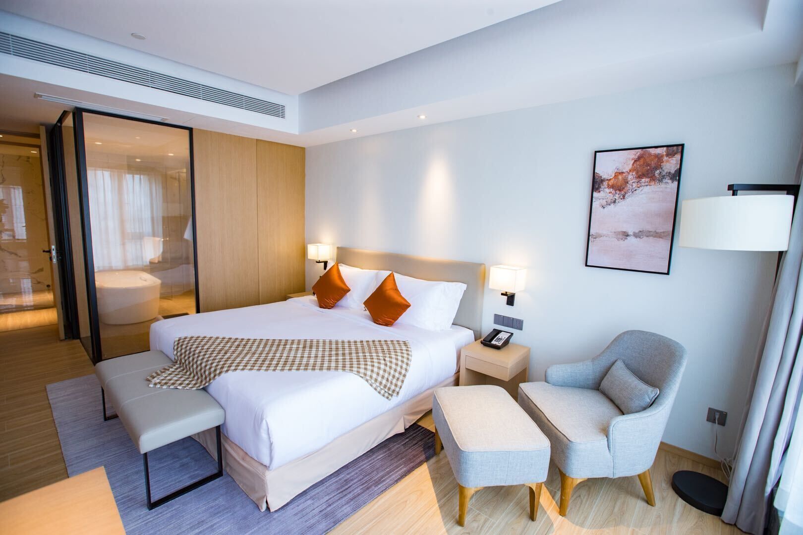 A modern hotel room with a large bed, two bedside tables with lamps, a grey armchair, and a small table.