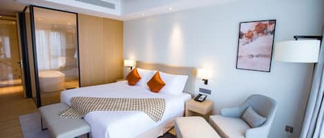 A modern hotel room with a large bed, two bedside tables with lamps, a grey armchair, and a small table.