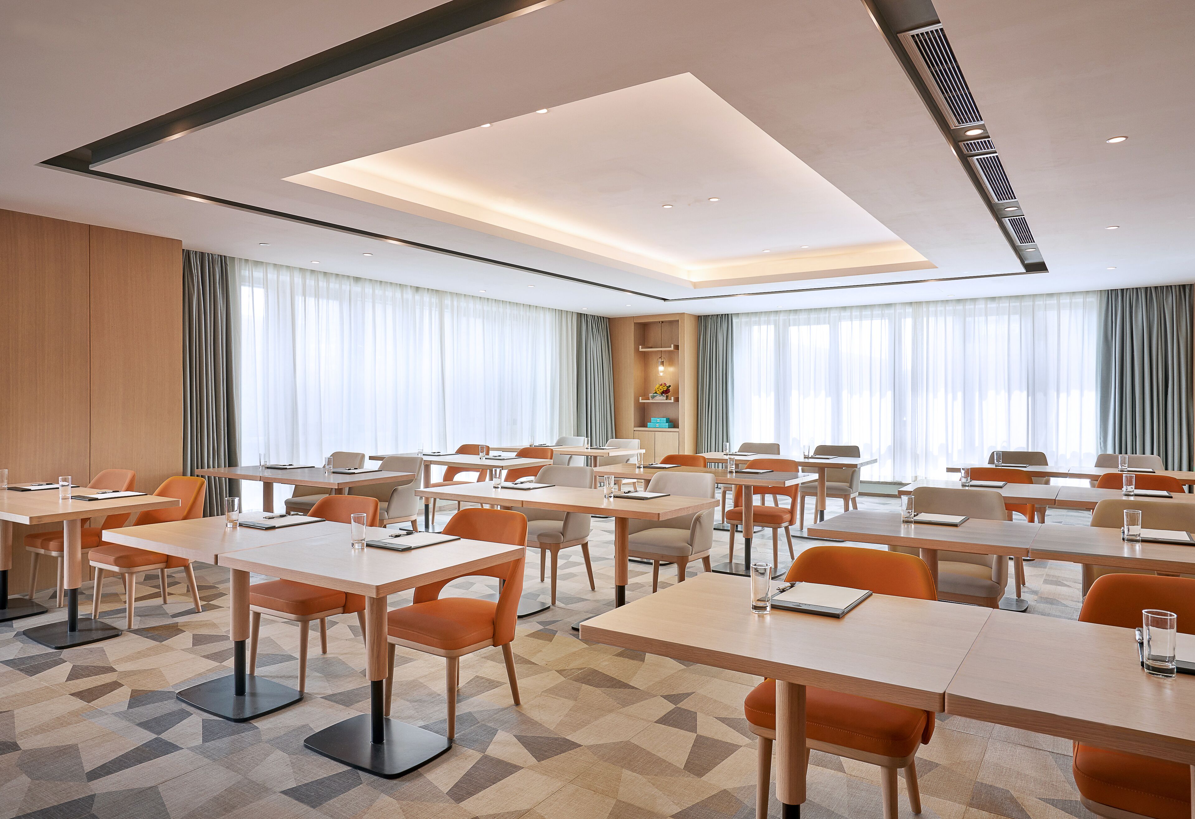 Un salón de conferencias con varias mesas y sillas, amplias ventanas con cortinas y un diseño moderno en el techo.