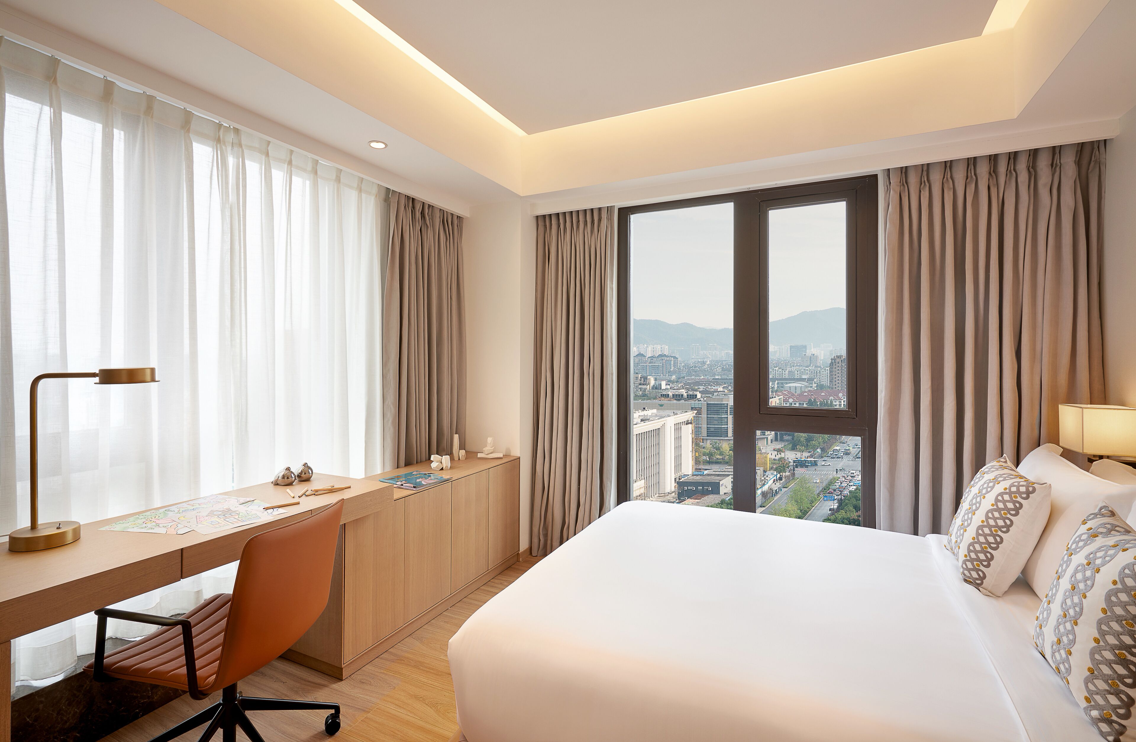 A hotel room with a large bed, a desk with a chair, a view of the city through large windows, and a cityscape visible outside.