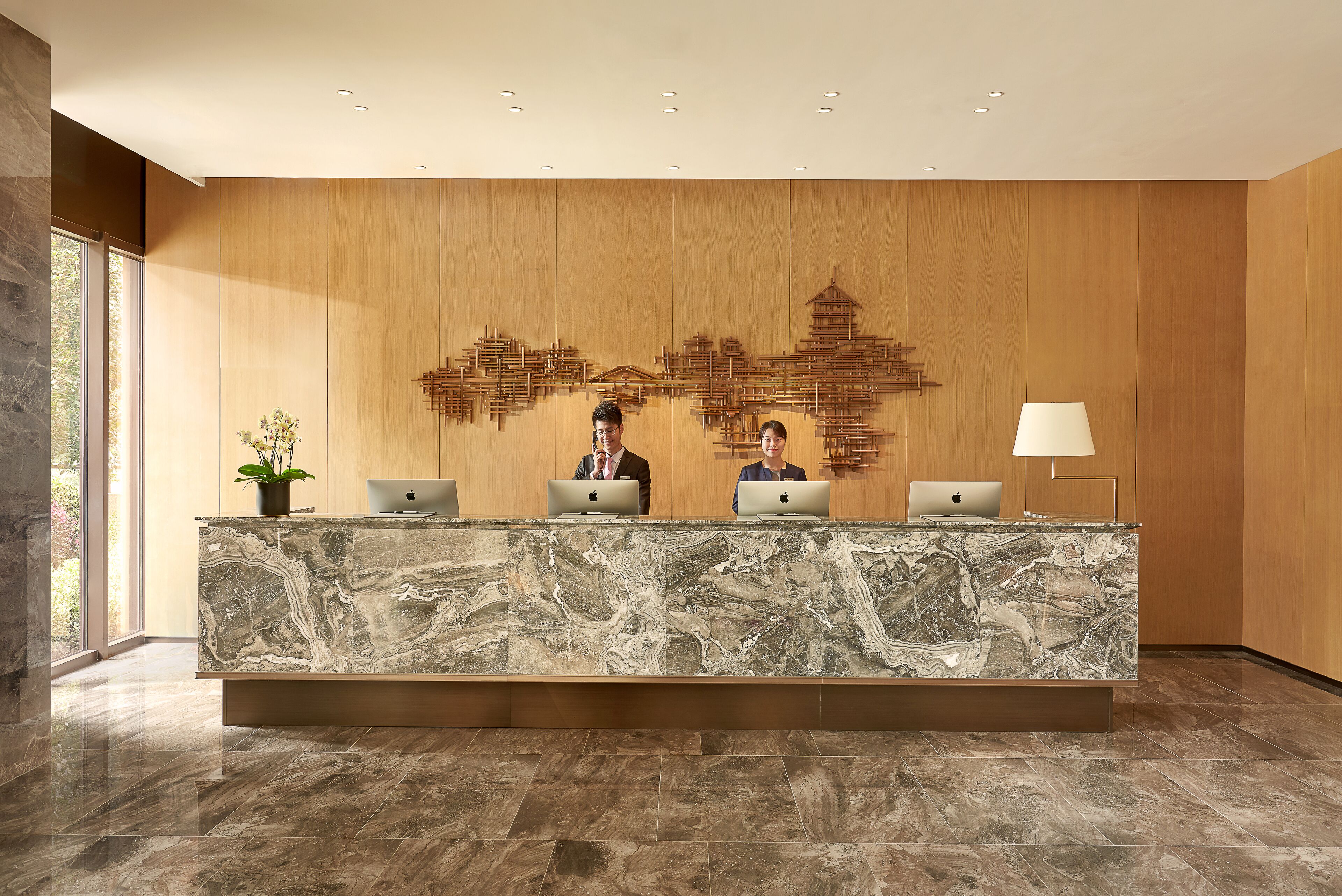 A reception desk with marble top, two people seated behind it, and a wooden wall with a cityscape mural.