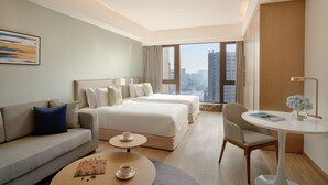 A hotel room with a sofa, two beds, a coffee table, a chair, and a view of the city.