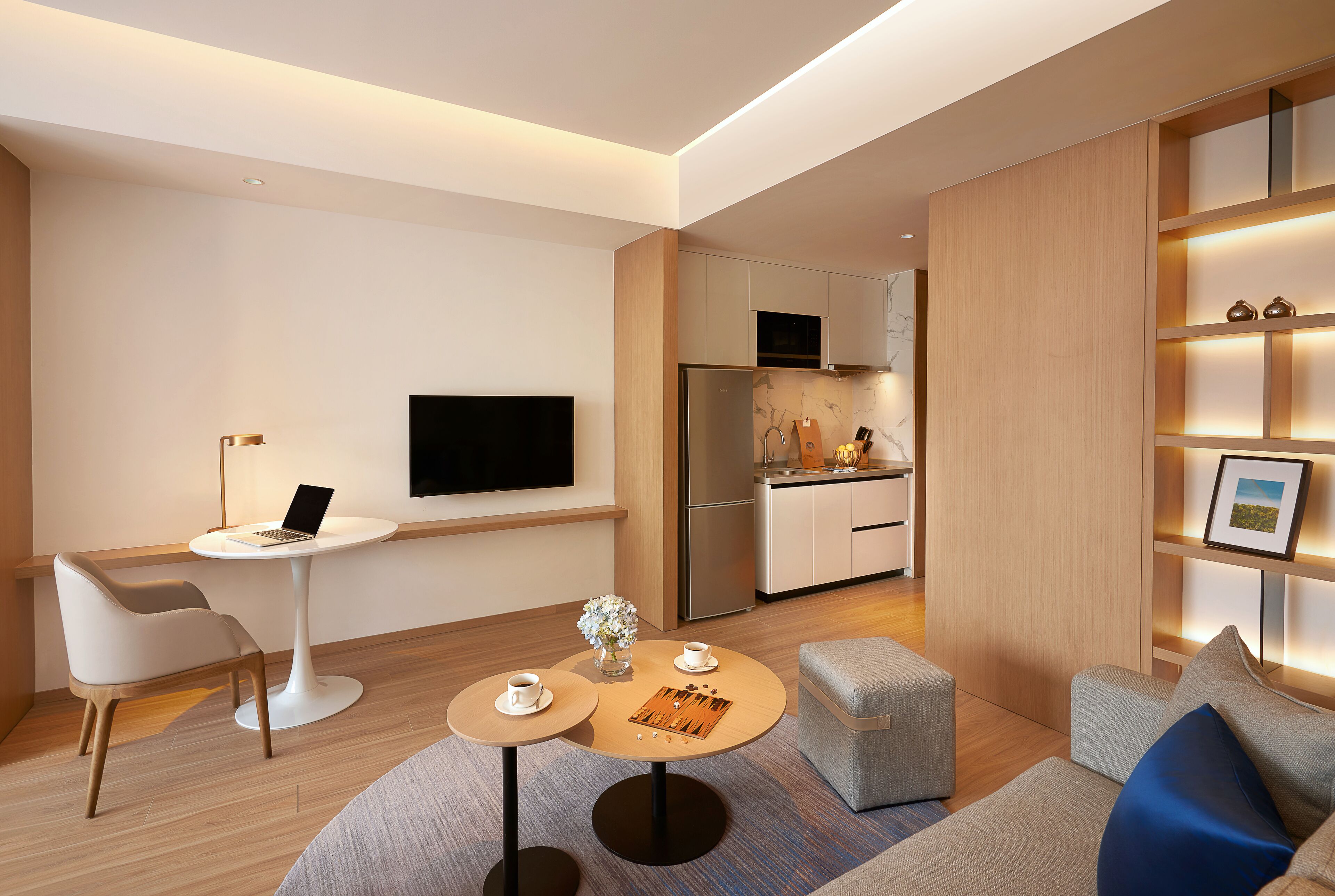 A modern living room with a flat-screen TV, a dining table, a sofa, and a kitchen area with a refrigerator and microwave.