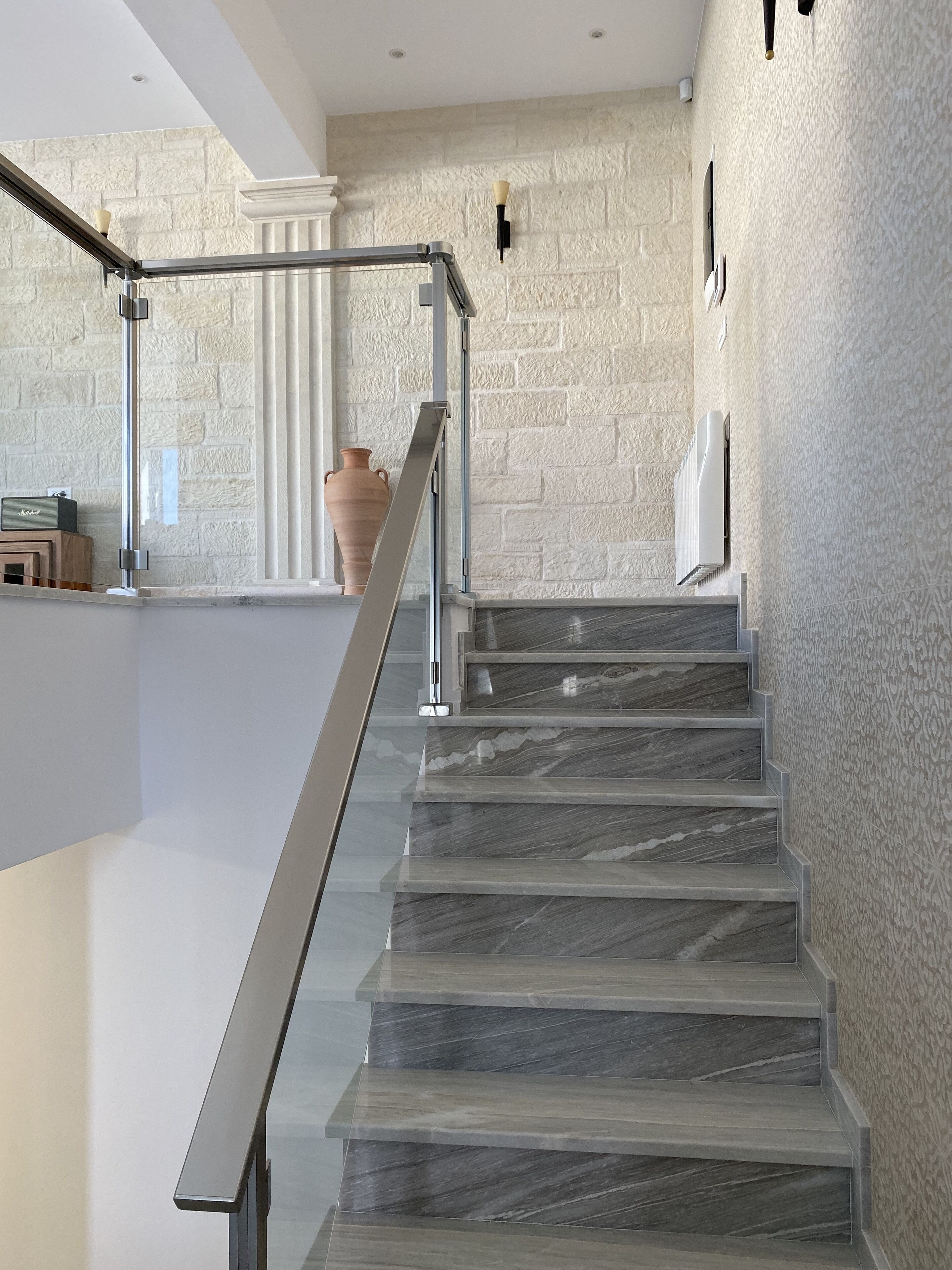 Un escalier moderne avec des marches en marbre et une rampe en métal, menant à une pièce dont un des murs est en pierre et qui contient une plante en pot.
