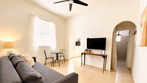 Un salon comprenant un canapé, un téléviseur posé sur un meuble, une table à manger avec des chaises et un ventilateur de plafond.