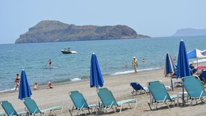 A beach with rows of blue and white lounge chairs, a few umbrellas, and a distant rocky island. - Villa Giorgos (Platanias)