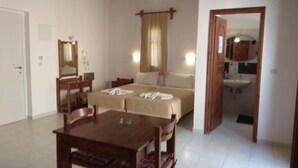 A room with a wooden dining table and chairs, a bed with a beige cover, a mirror, and a small kitchenette with a sink. - Villa Giorgos (Platanias)