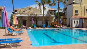 A swimming pool with lounge chairs and umbrellas, surrounded by palm trees and buildings. - Villa Giorgos (Platanias)