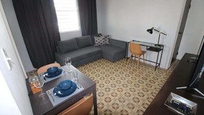 A compact living space with a dining table set for two, a sofa, a desk with a lamp, and a television. - A2-Quaint 1B-Fast WiFi-Walk to Restaurants, Beach (San Juan)