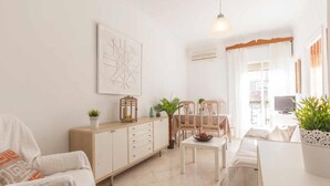 A bright living room with a white sofa, a wooden cabinet, and a dining table with chairs. There is a hanging light fixture and a painting on the wall. - Beautiful apt 2 rooms Cathedral views cn WIFI (Sevilla)