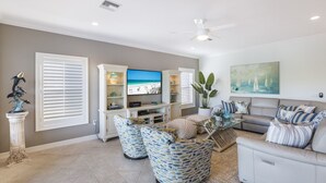 A living room with a flat-screen TV, a ceiling fan, and a large window with blinds. - Beach Bay Serenity: Pine Ave canal front pool home (Anna Maria)