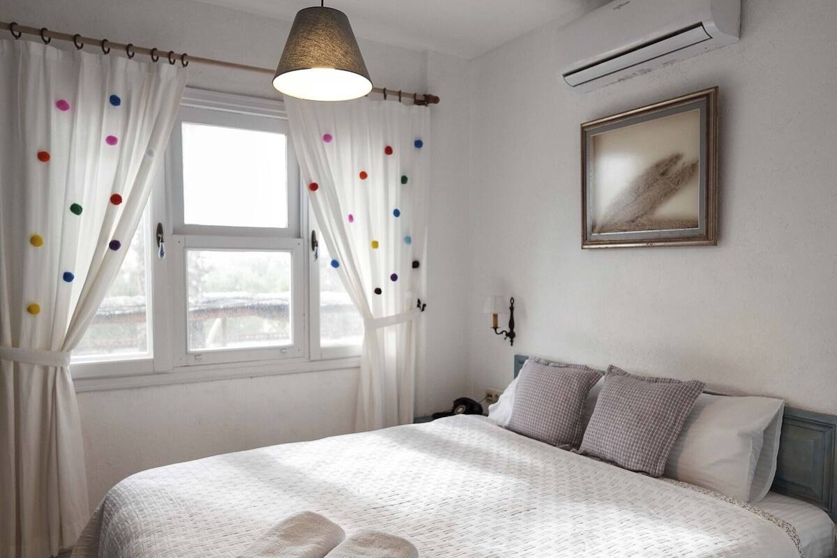 A bedroom with a bed, a hanging light, a window with curtains, an air conditioner, and a painting on the wall.