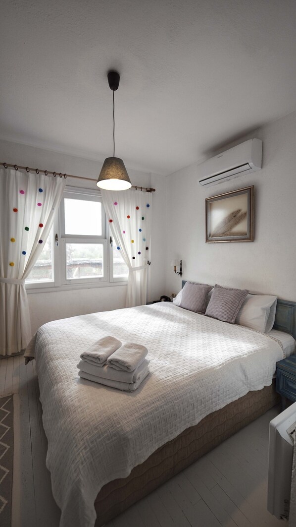 Un dormitorio con cama, lámpara colgante, ventana con cortinas, aire acondicionado y un cuadro en la pared.