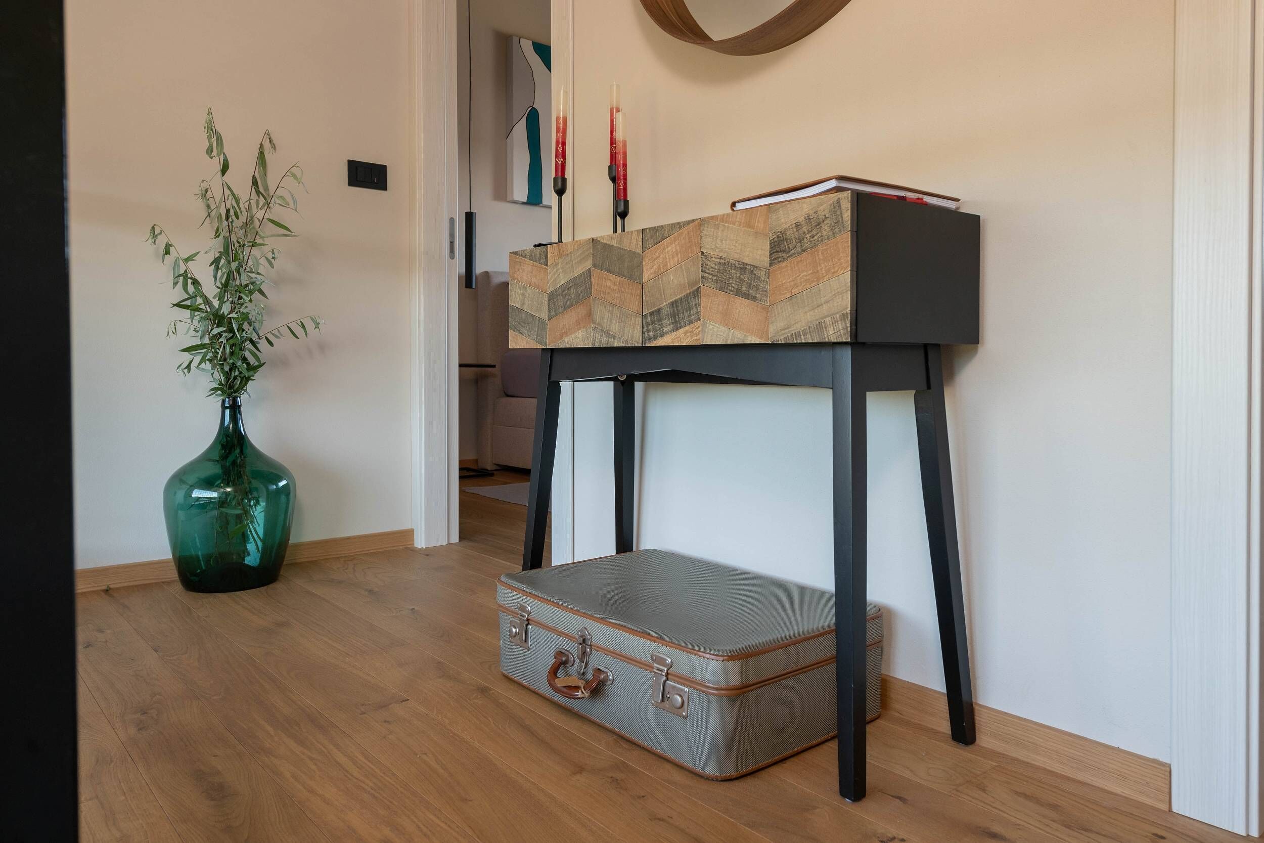 A room with a wooden floor, a green vase with a plant, a black and patterned table, and a suitcase on the floor.