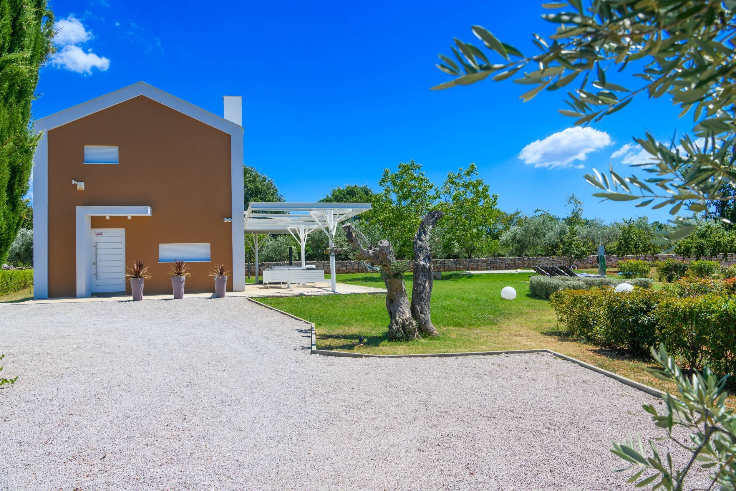 Um edifício moderno com fachada marrom, área de estar externa coberta e entrada com caminho de cascalho.