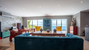 A spacious living room with a large teal sofa, a wooden coffee table, and a view of the ocean through large windows. - Mansão Macau by LovelyStay (Arco Da Calheta)
