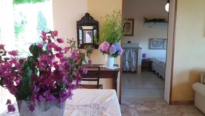 Una habitación con una mesa de comedor, sillas, mantel blanco, un florero con flores moradas, un espejo y una cama con cubrecama blanco.