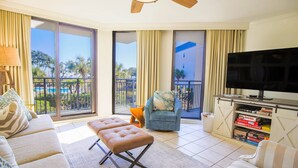 A spacious living room with a large TV, comfortable seating, a balcony with a pool view, and a sliding door leading to an outdoor area.