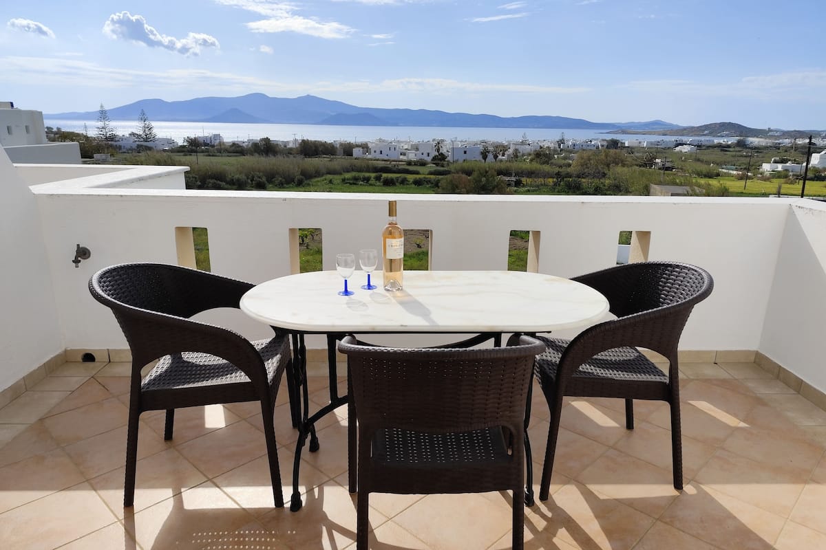 A round table with four wicker chairs on a terrace with a view of the sea and mountains.