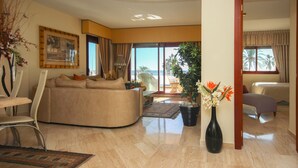A spacious living area with a curved sofa, a dining table with chairs, a large vase with flowers, and a view of the beach through large windows. - First floor Beach side property, San Pedro (Marbella)
