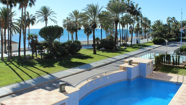 A swimming pool with a curved design, surrounded by palm trees and a view of the sea. - First floor Beach side property, San Pedro (Marbella)