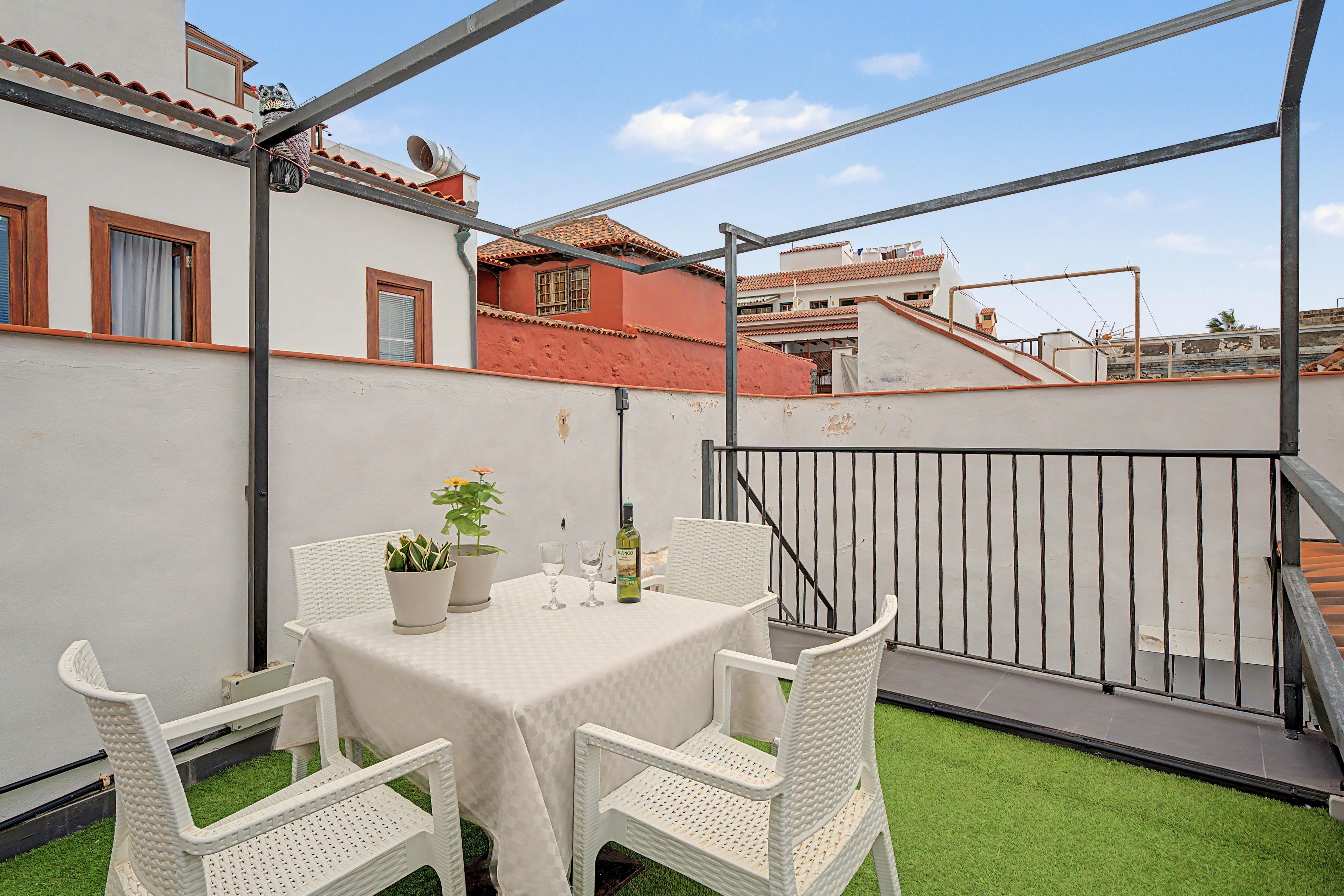 Terraza en la azotea con una mesa para dos, rodeada de sillas de mimbre blanco, una planta en maceta y una botella de vino.