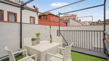 Terraza en la azotea con una mesa para dos, rodeada de sillas de mimbre blanco, una planta en maceta y una botella de vino.