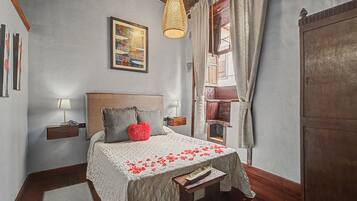 Un dormitorio con cama, mesita de noche, lámpara, lámpara colgante, ventana con cortinas y puerta.