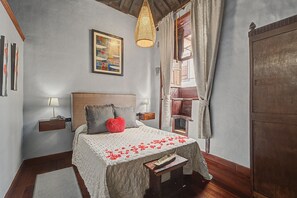 Ein Schlafzimmer mit einem Bett, einem Nachttisch, einer Lampe, einer Hängeleuchte, einem Fenster mit Vorhängen und einer Tür.