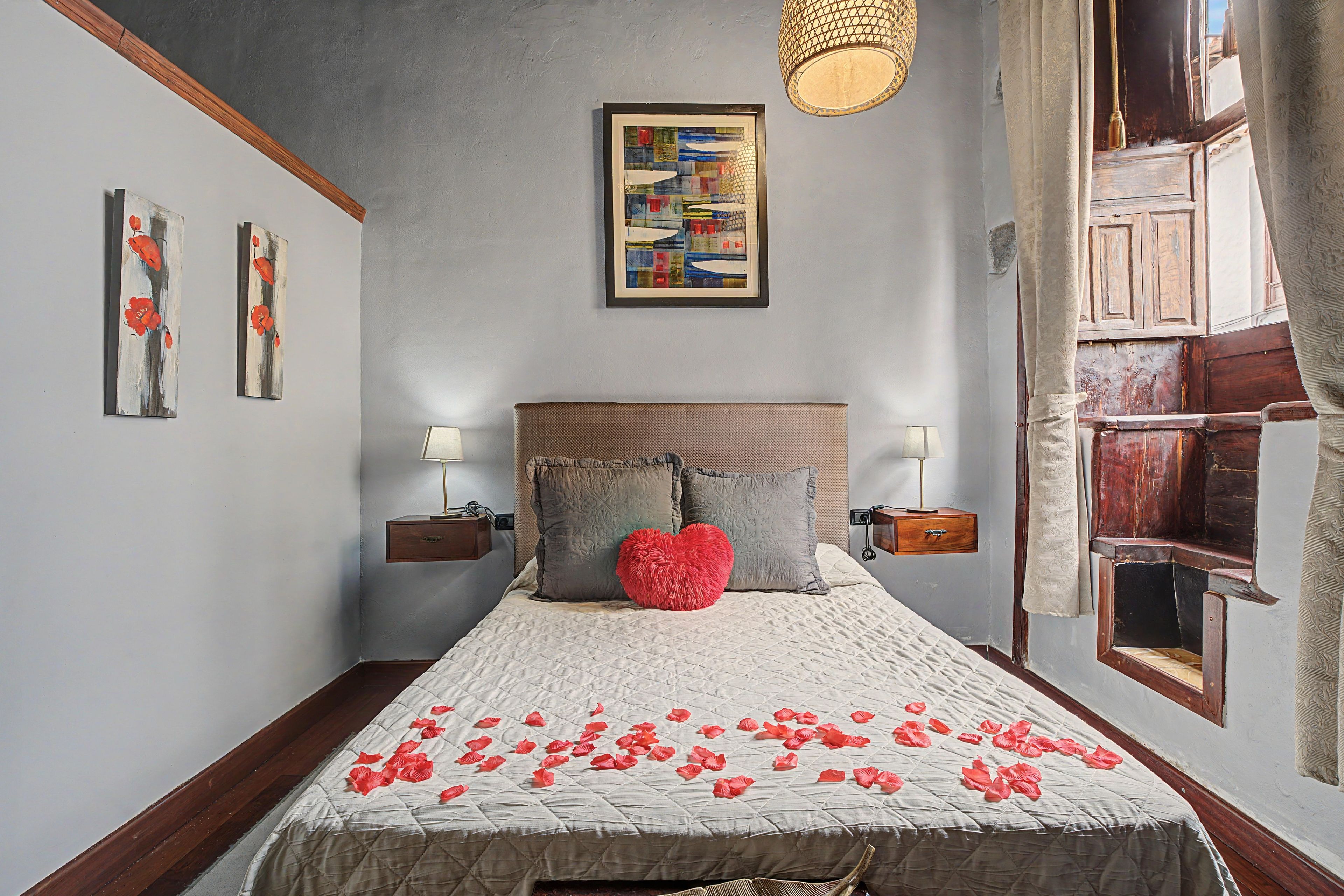 A bedroom with a bed, two bedside tables, a nightstand, a wall-mounted lamp, and a window with curtains.