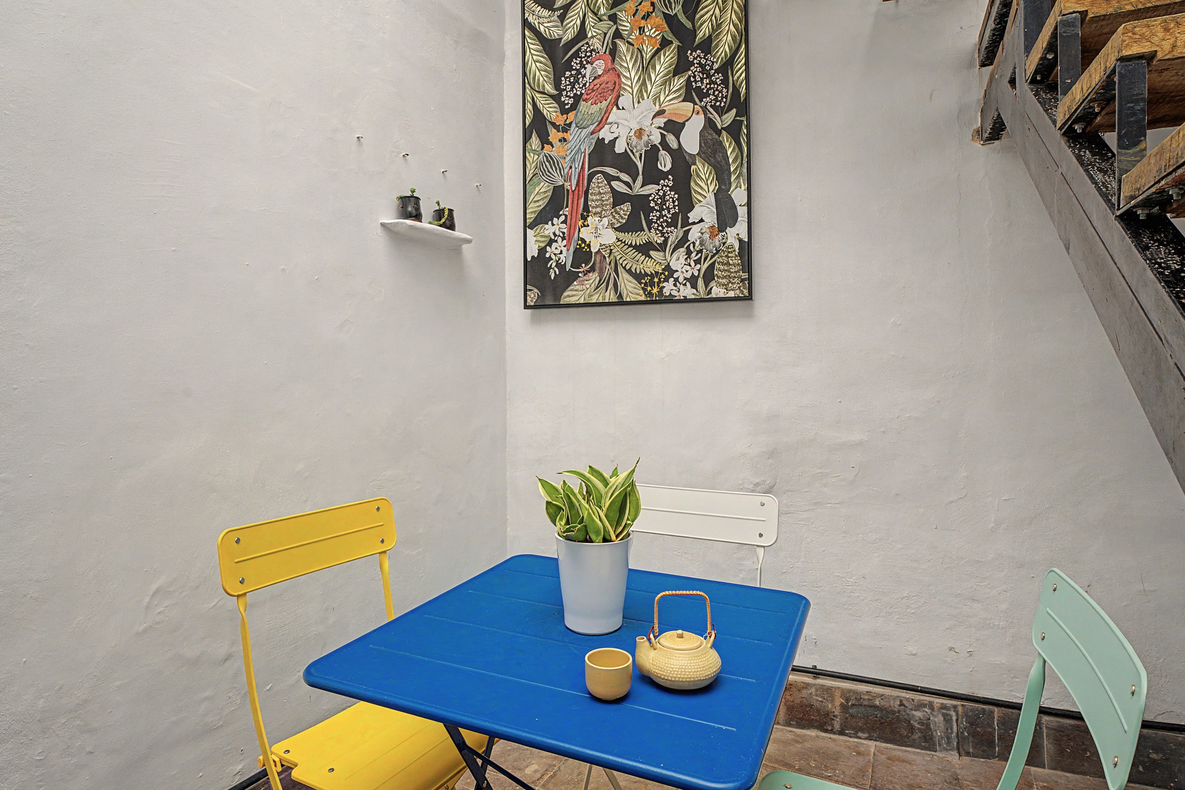 A small outdoor seating area with a blue table, yellow and green chairs, a potted plant, and a teapot.